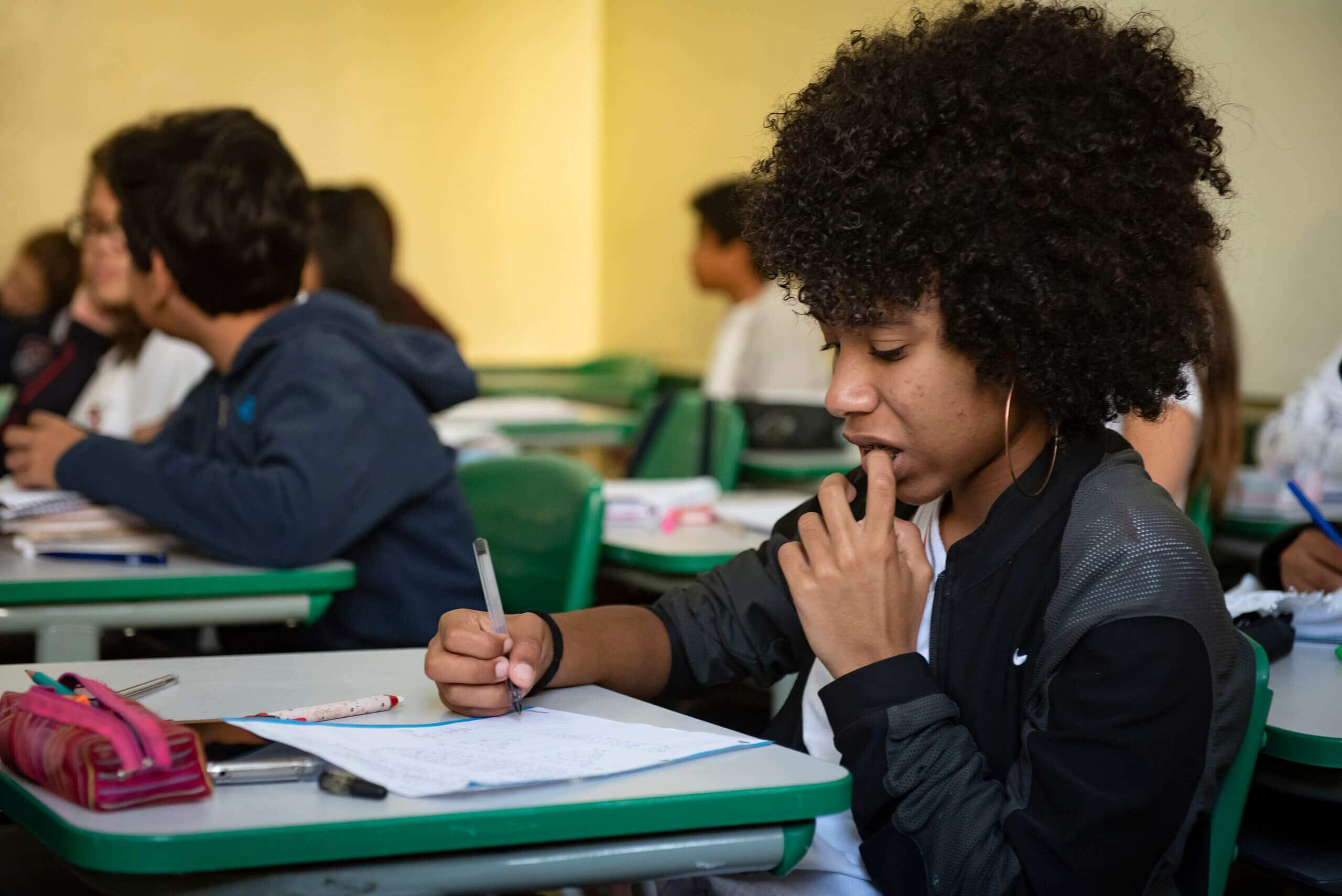 Aluna adolescente (aproximadamente 14 anos) escrevendo em uma folha pautada com cara apreensiva ou nervosa sentada em uma carteira na sala de aula. Ela é negra, tem cabelo black power, usa um brinco de argolas douradas e um casaco preto por cima da blusa branca do uniforme da escola. Ao fundo outros alunos aparecem também sentados nas carteiras.